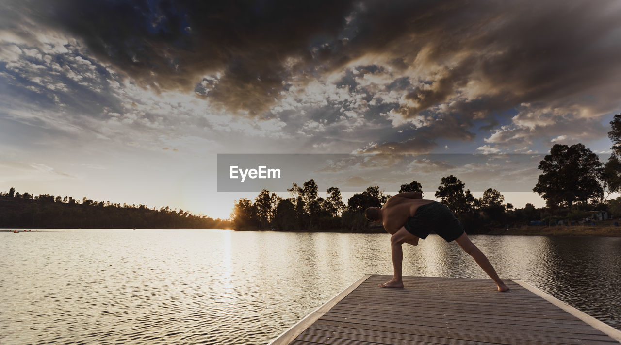 Shirtless man practicing yoga on pier against sky during sunset