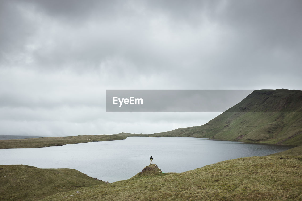 Distant view of man standing by lake against cloudy sky