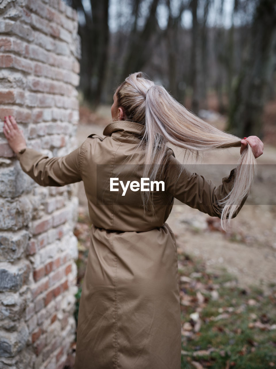 Rear view of a young woman in a beige coat outside leaning against a brick wall with her left hand and stretching her long blond hair with her right hand Woman Back Hair Blond Beige Outdoor Hand Bricks Trees Body No Face