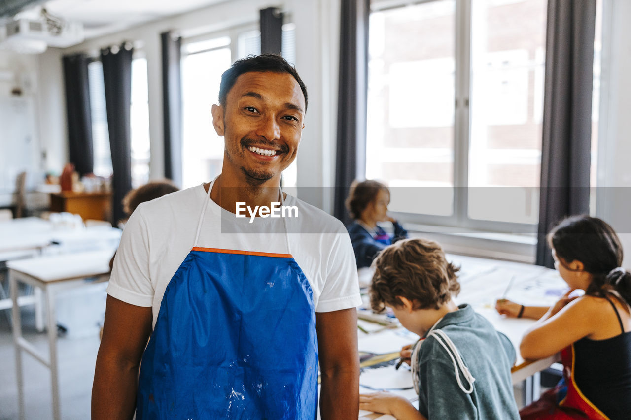 Portrait of smiling male art teacher wearing blue apron in classroom at school