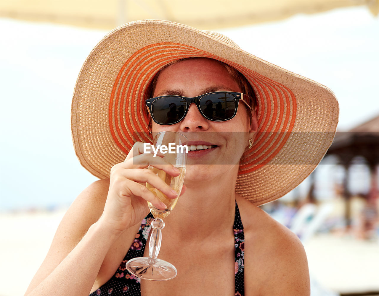 Portrait of woman in sun hat and sunglasses holding champagne at beach