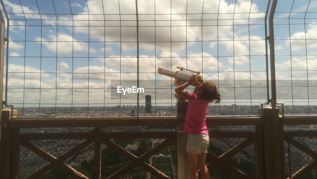 Girl looking through telescope by fence against cloudy sky