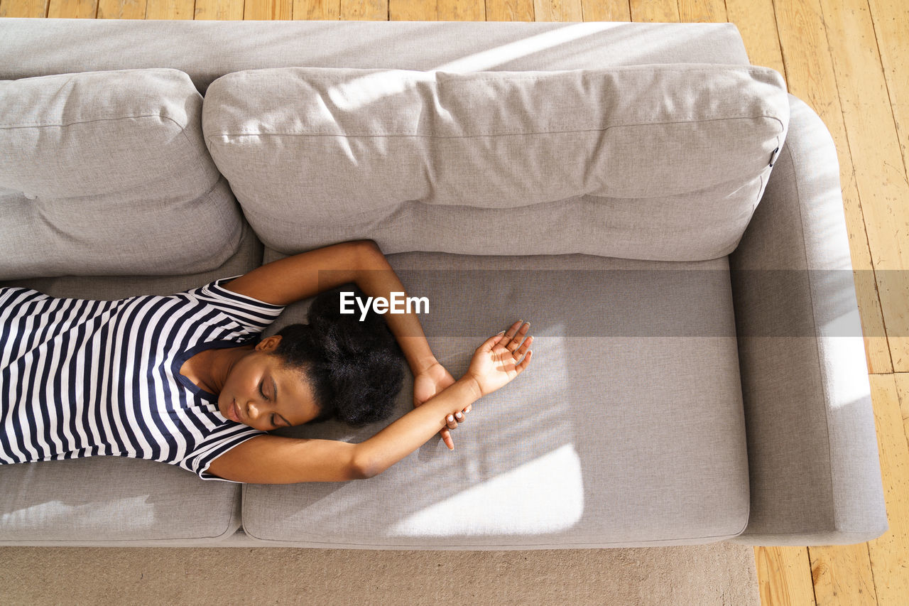 African american young woman wear stripped t-shirt resting, sleeping on couch with arms up at home