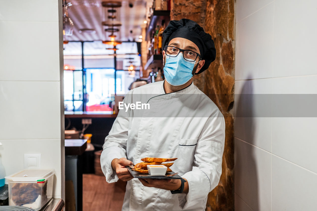 Serious male chef in uniform and medical mask standing with delicious snacks on slate board in cafe and looking at camera