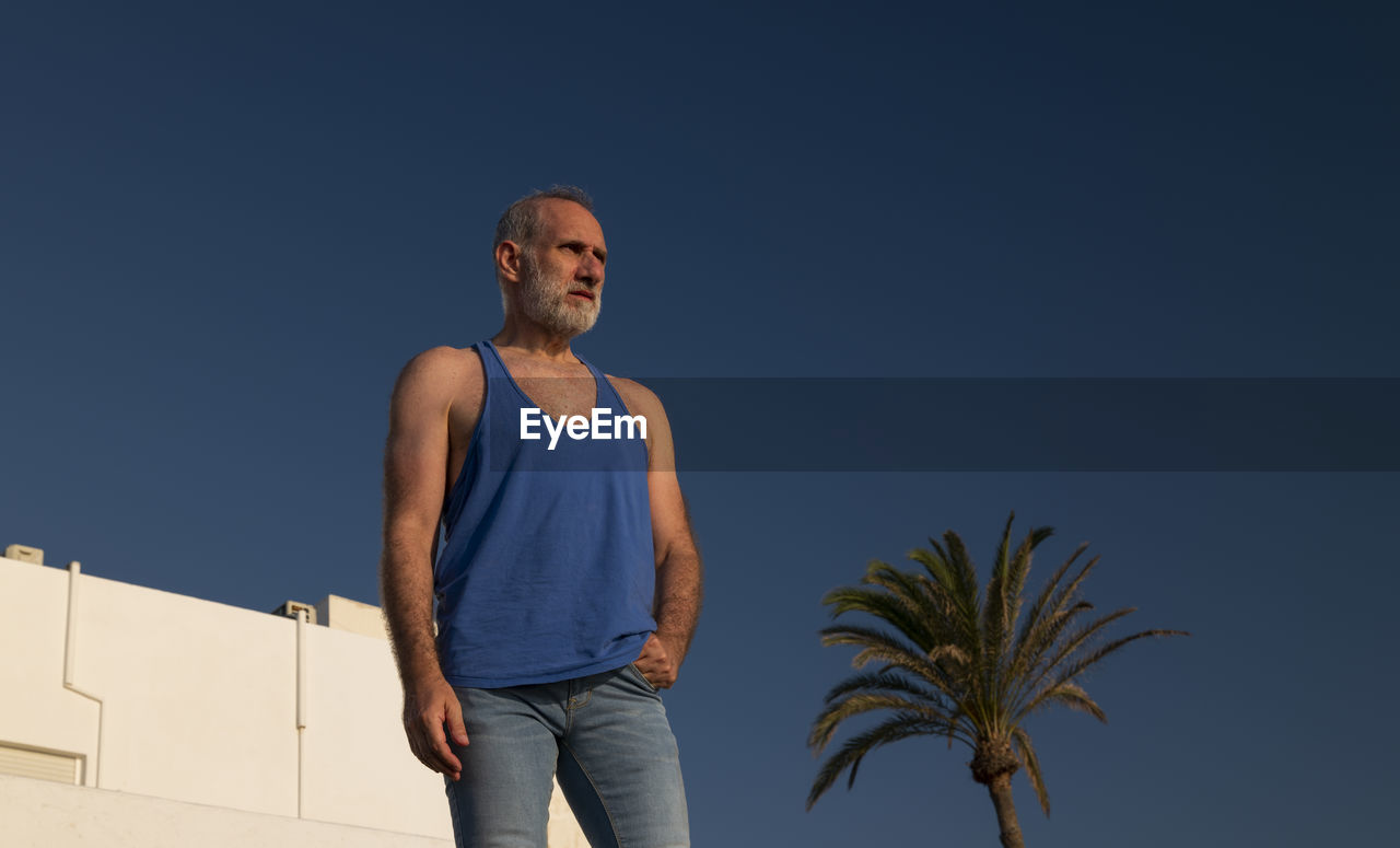 Portrait of adult man looking away against white building, palm tree and blue sky in summer