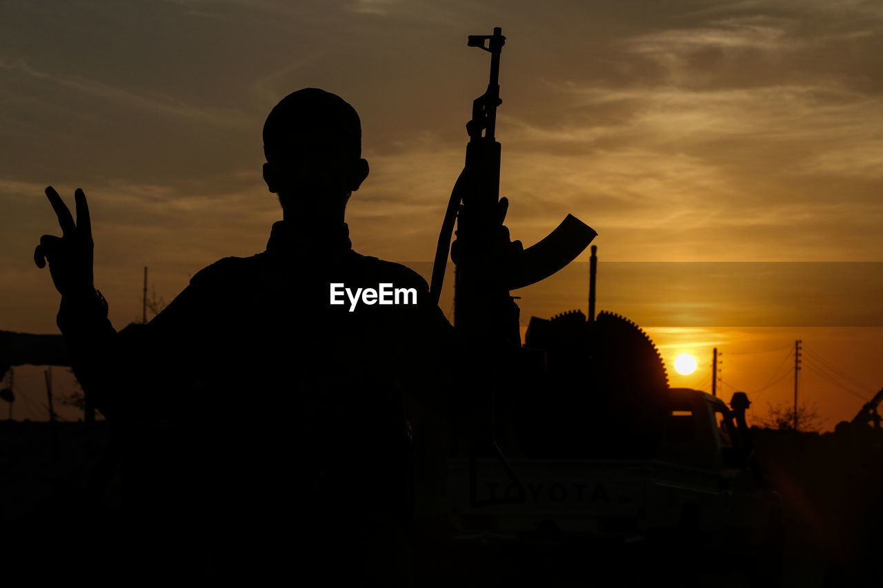 Silhouette soldier holding rifle standing against sky during sunset