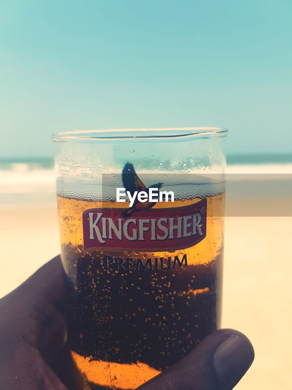 CLOSE-UP OF HAND HOLDING DRINK AGAINST SEA