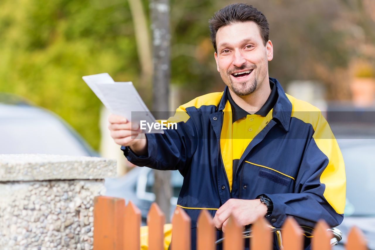 Portrait of smiling postal worker holding yellow while standing outdoors