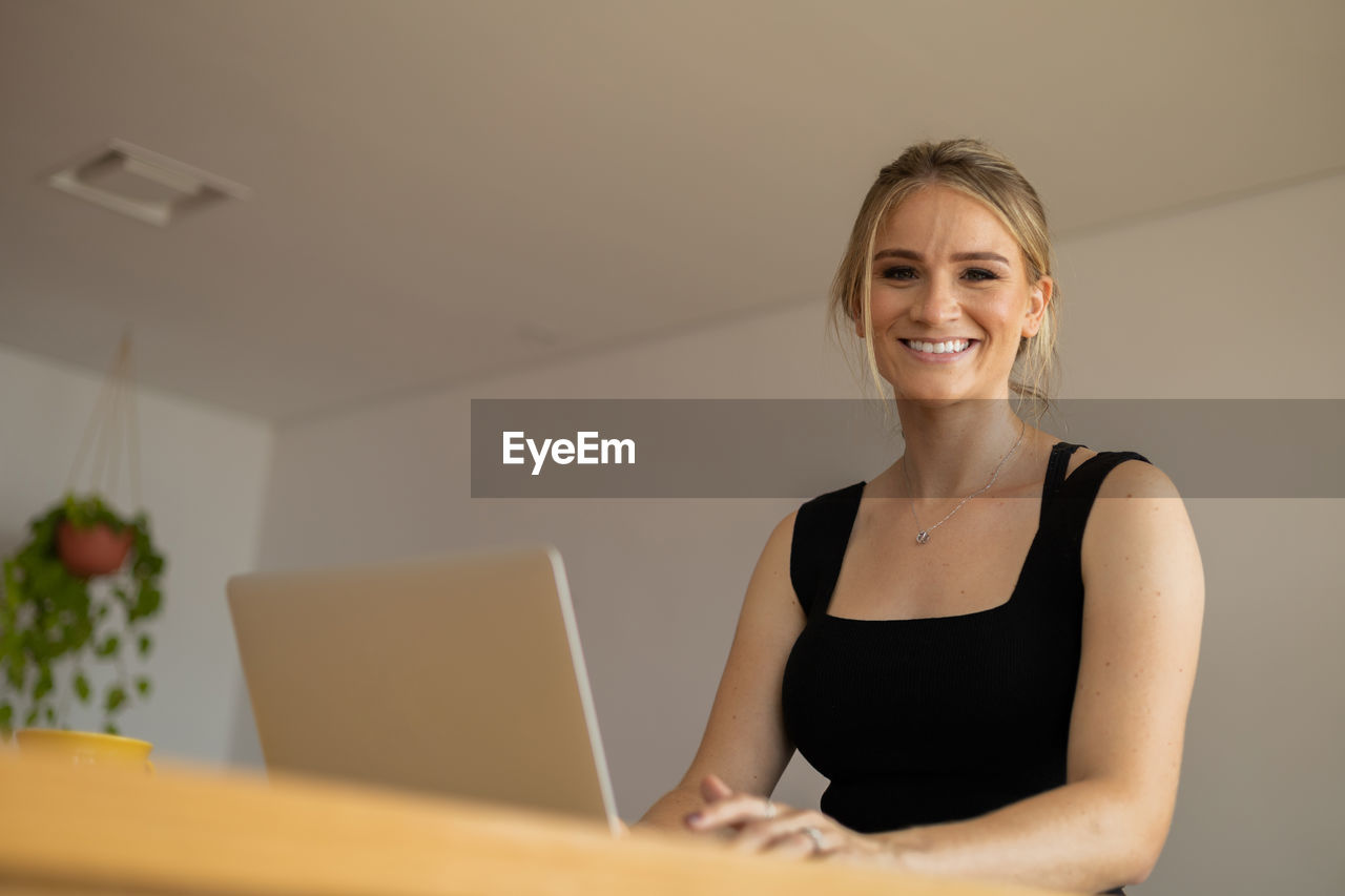 smiling, adult, one person, women, computer, laptop, happiness, portrait, indoors, wireless technology, technology, business, using laptop, businesswoman, looking at camera, working, emotion, cheerful, office, female, business finance and industry, communication, blond hair, person, copy space, occupation, lifestyles, table, internet, front view, sitting, computer network, clothing, smile, teeth, waist up, corporate business, human face, room, entrepreneur, businesswear, young adult, hairstyle, professional occupation