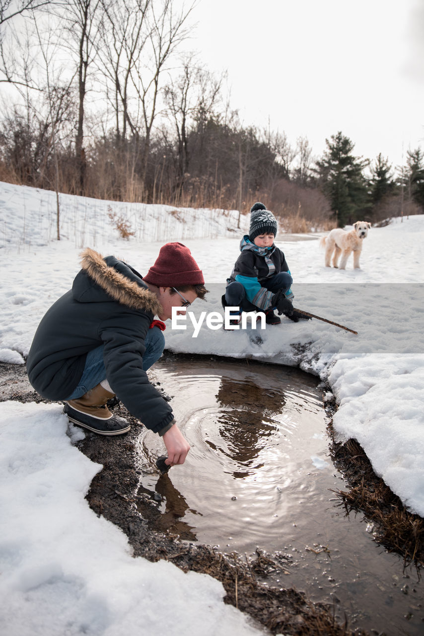 Two boys playing in a puddle on a snowy path with dog in background.