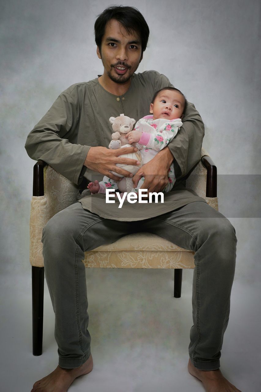 Portrait of father with daughter sitting on chair against wall