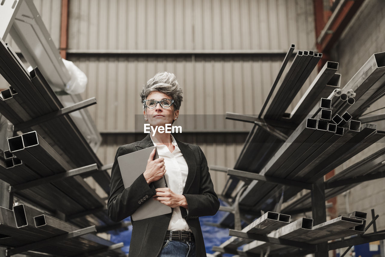 Mature businesswoman with laptop standing amidst metal beams on racks