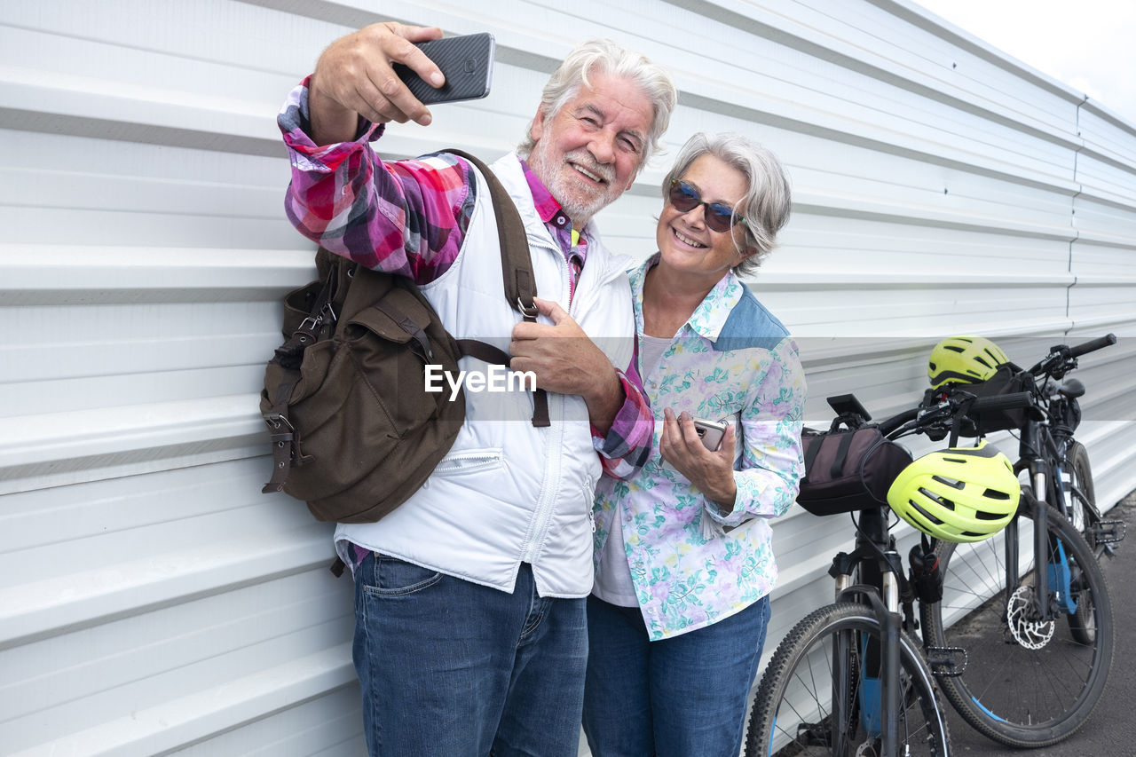 Smiling couple taking selfie while standing by wall outdoors