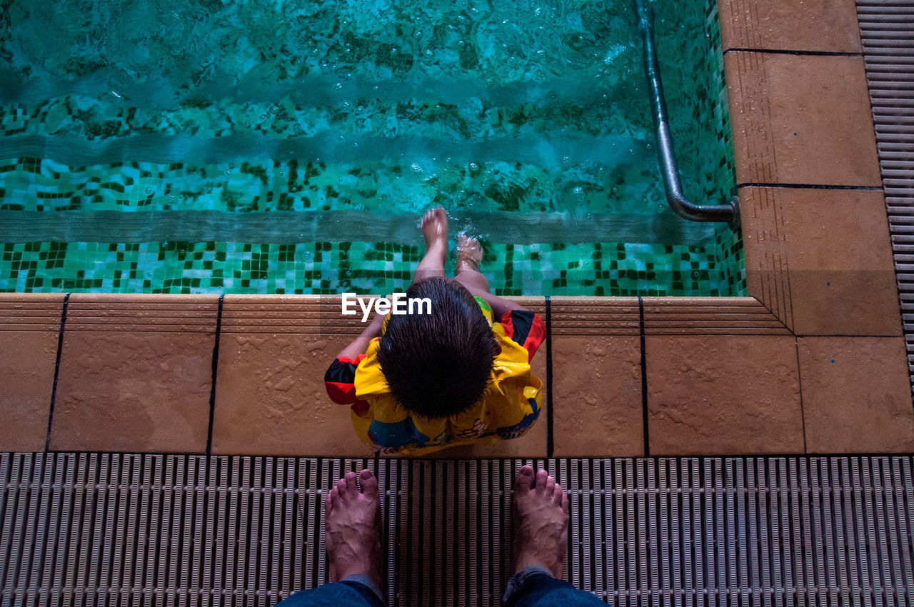 Low section of father standing by son sitting at poolside