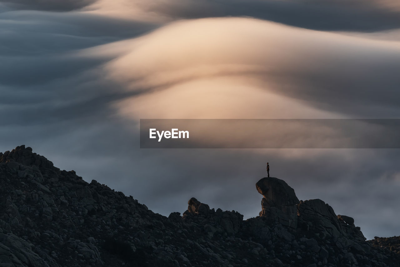 Silhouette of unrecognizable person standing on peak of rough cliff with colorful clouds on background