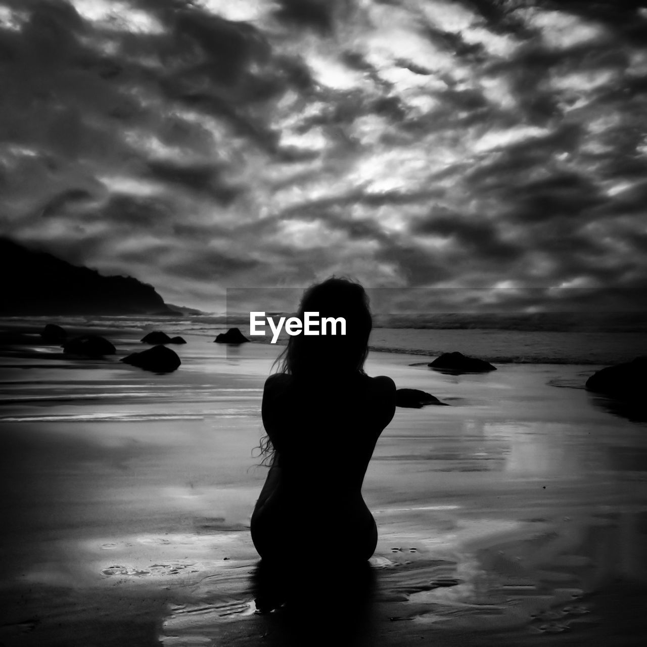 Rear view of woman sitting at beach against cloudy sky