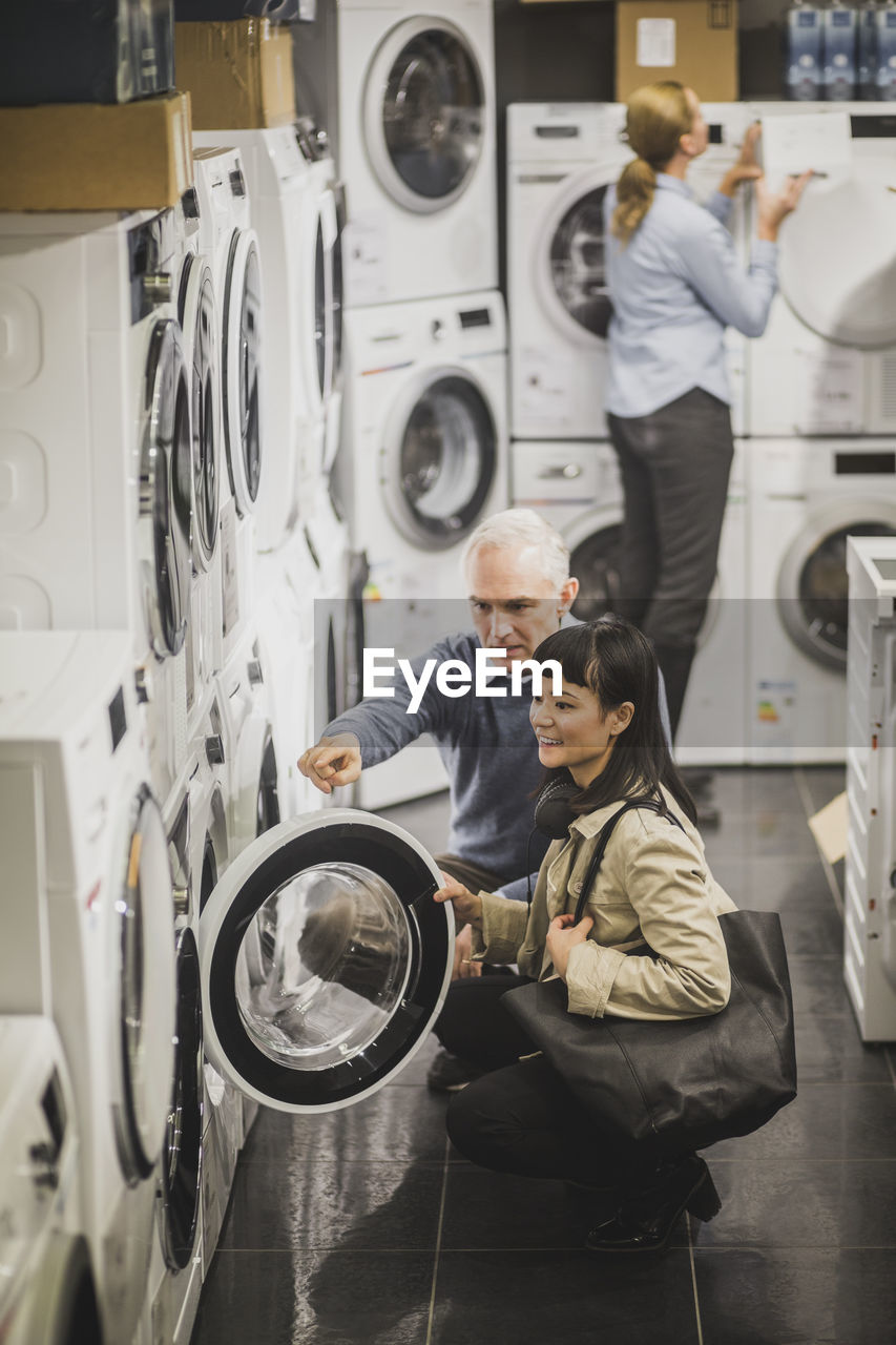 High angle view of mature owner pointing to washing machine while female customer crouching in electronics store