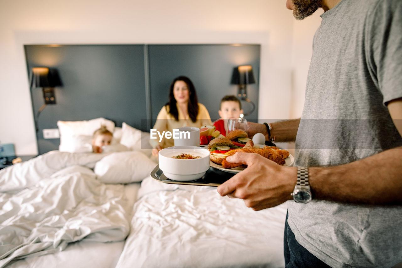 Father holding breakfast with family sitting on bed in background