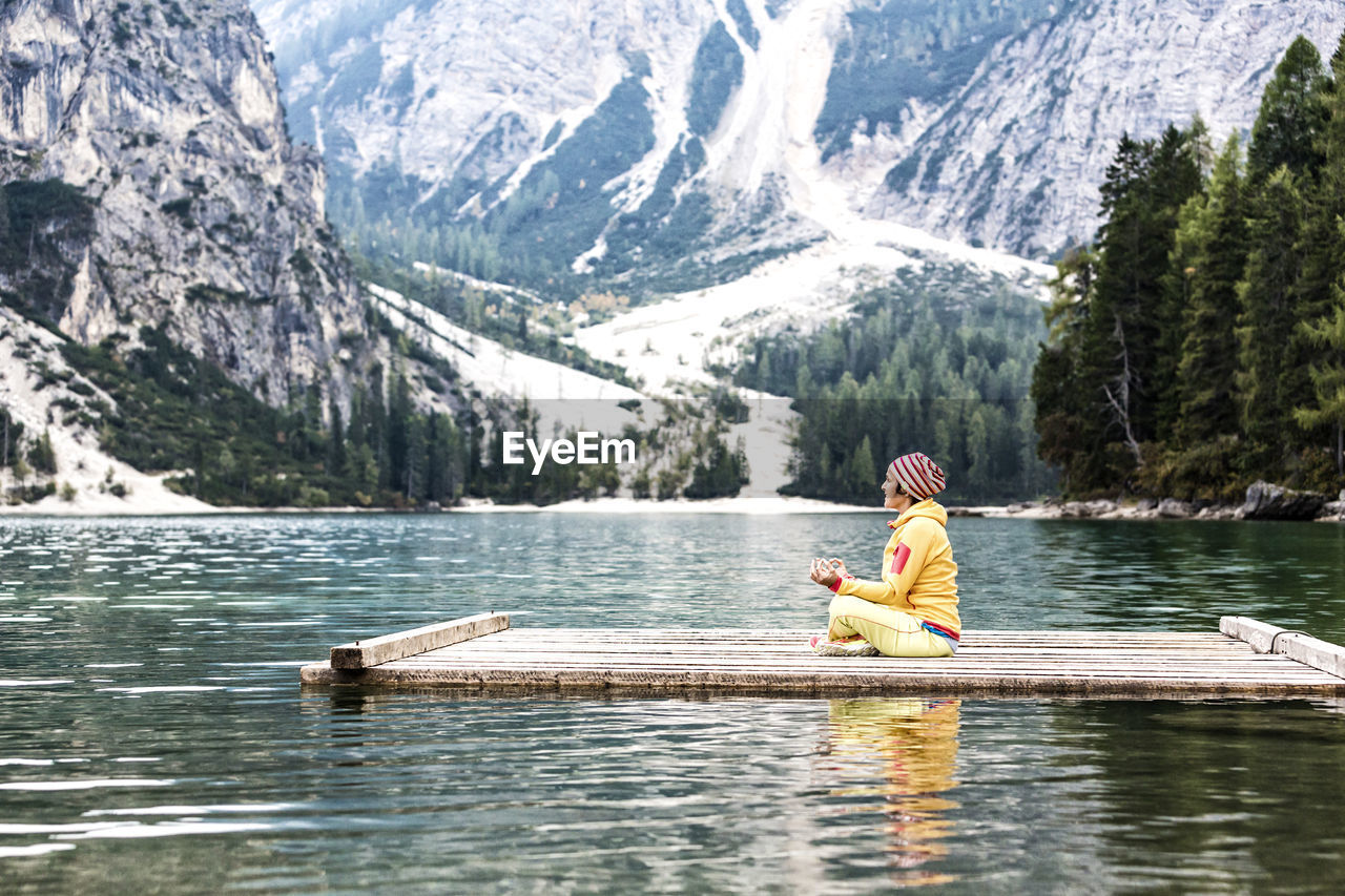 MAN SITTING ON LAKE AGAINST MOUNTAINS