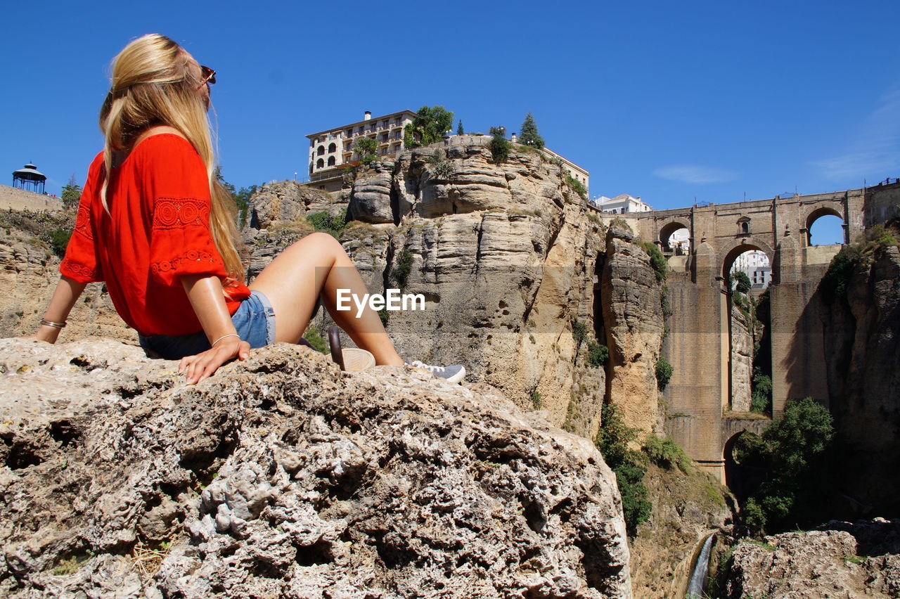 MIDSECTION OF WOMAN SITTING ON ROCK AGAINST SKY