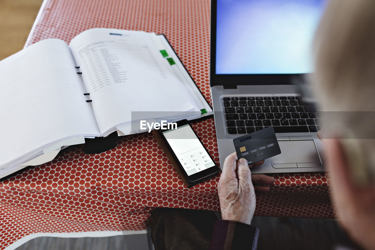 High angle view of retired senior man paying bills through credit card while using laptop at dining table