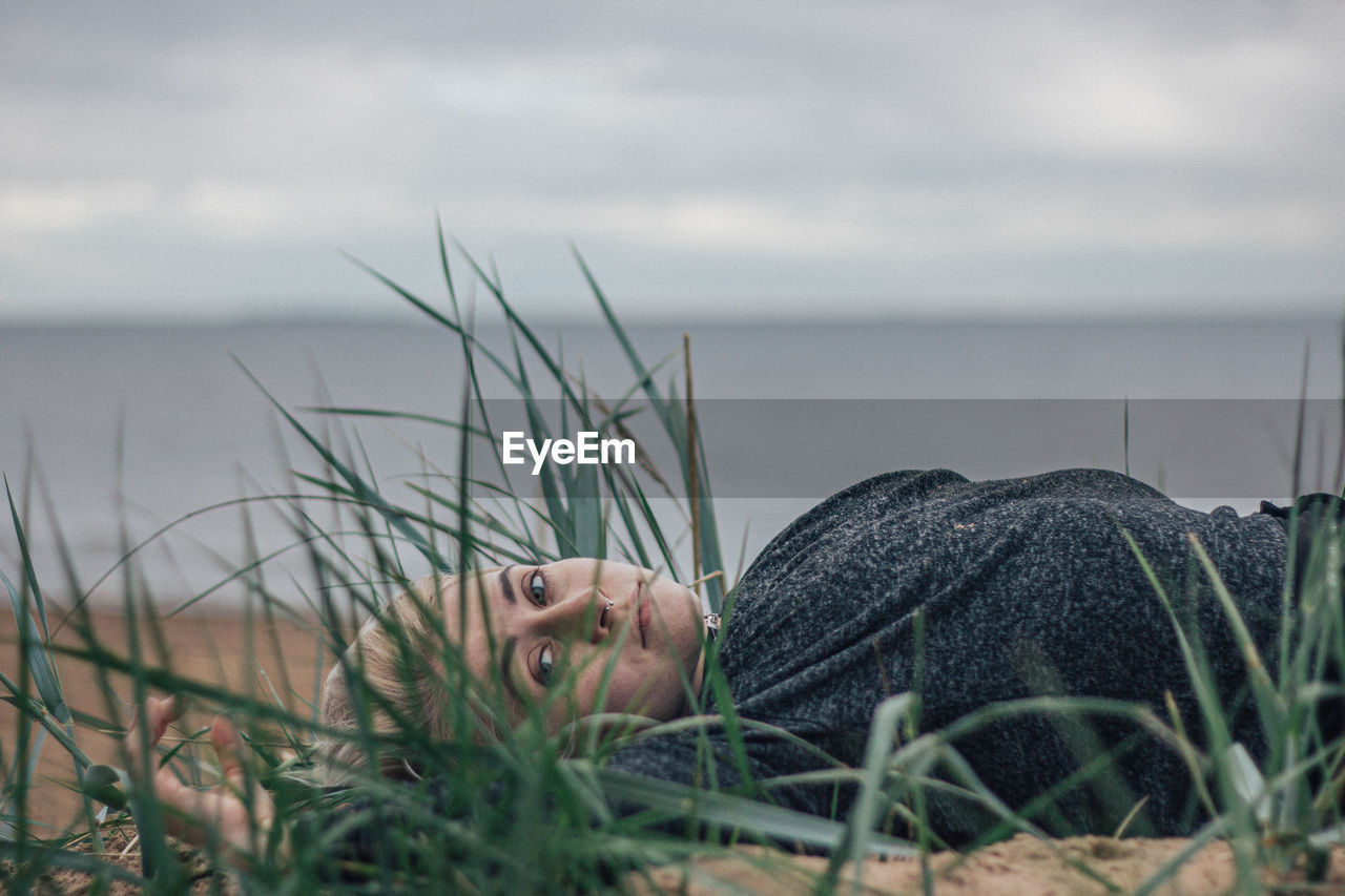 Portrait of young woman relaxing on sand at beach against sky