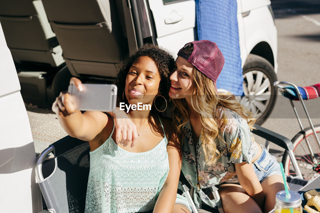 Cheerful women enjoying drink and nachos while sitting out of van in camping taking selfie with smartphone