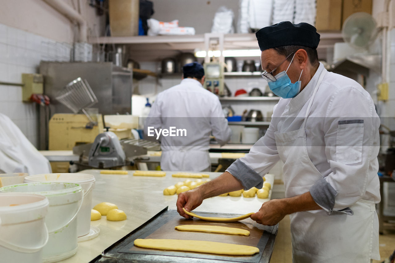 Side view of mature male cook in medical mask preparing raw dough for baking tasty pastry in bakehouse during coronavirus