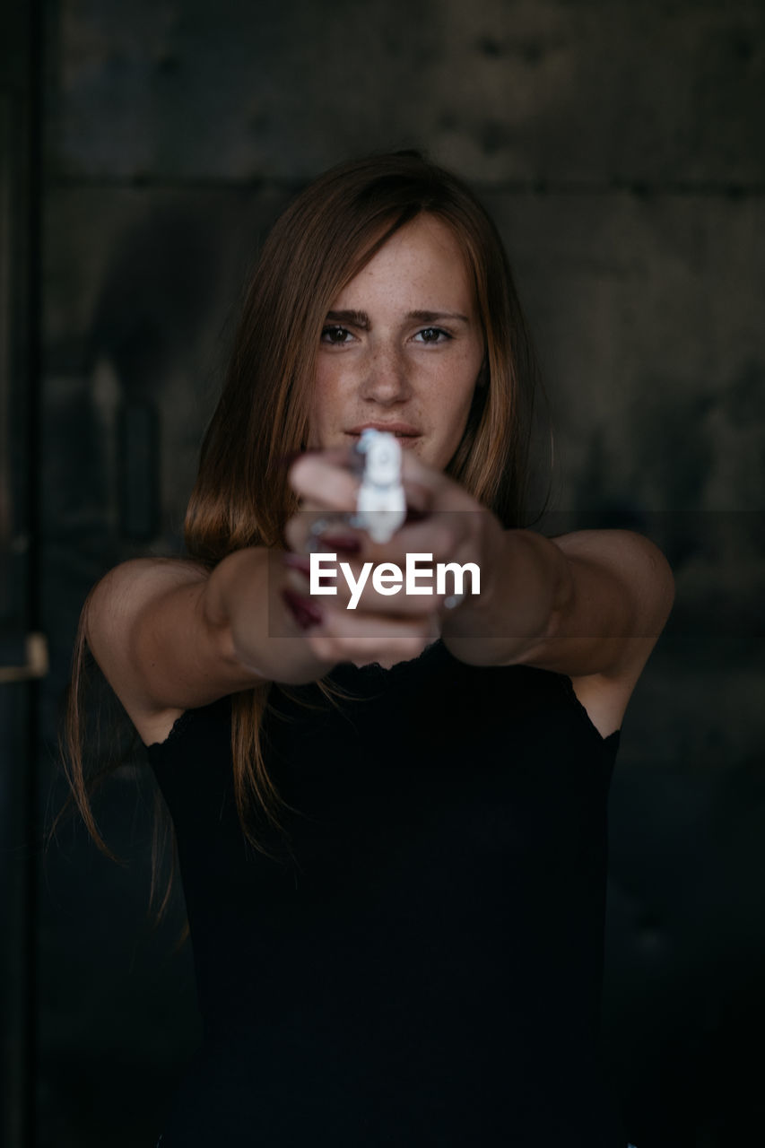 Portrait of young woman aiming gun while standing against wall