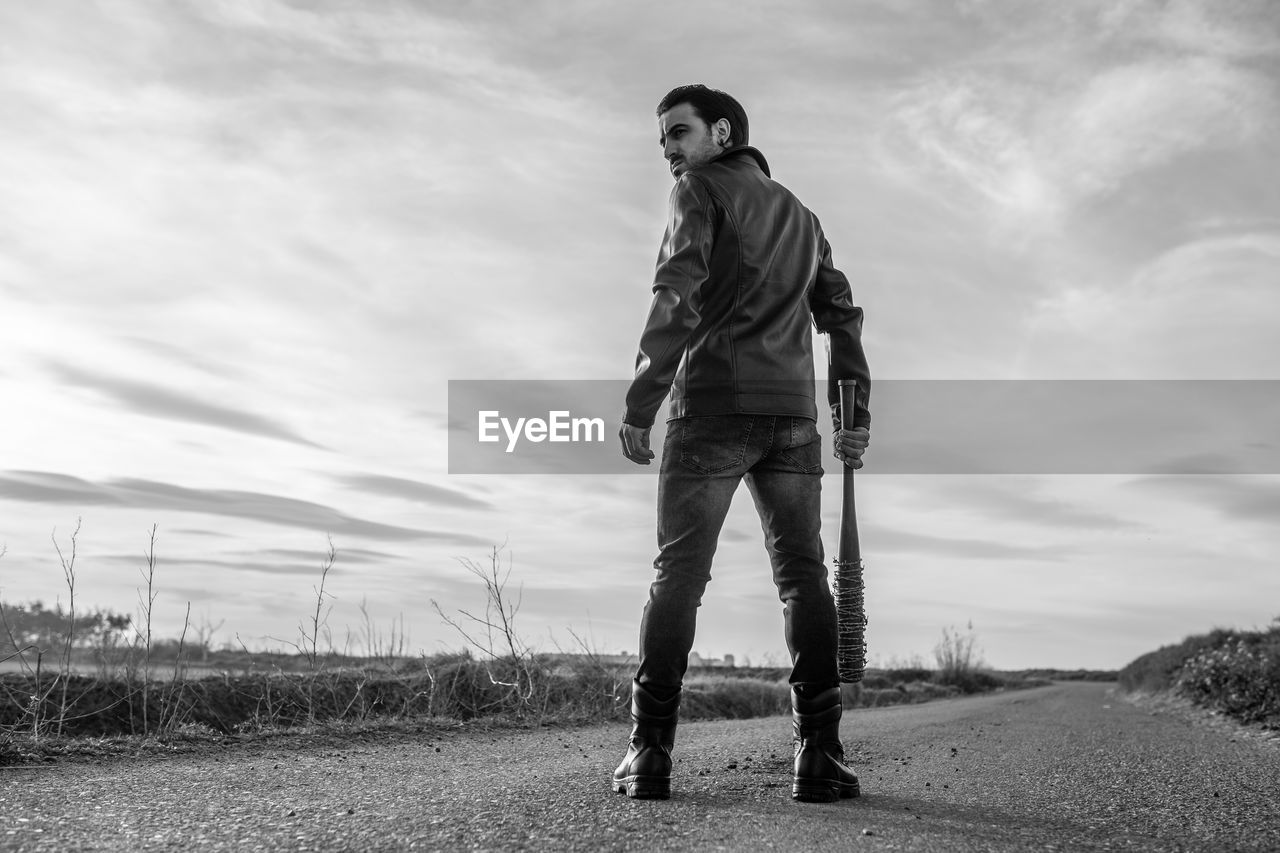 Rear view of mid adult man holding barbed wire wrapped baseball bat while standing on road against sky