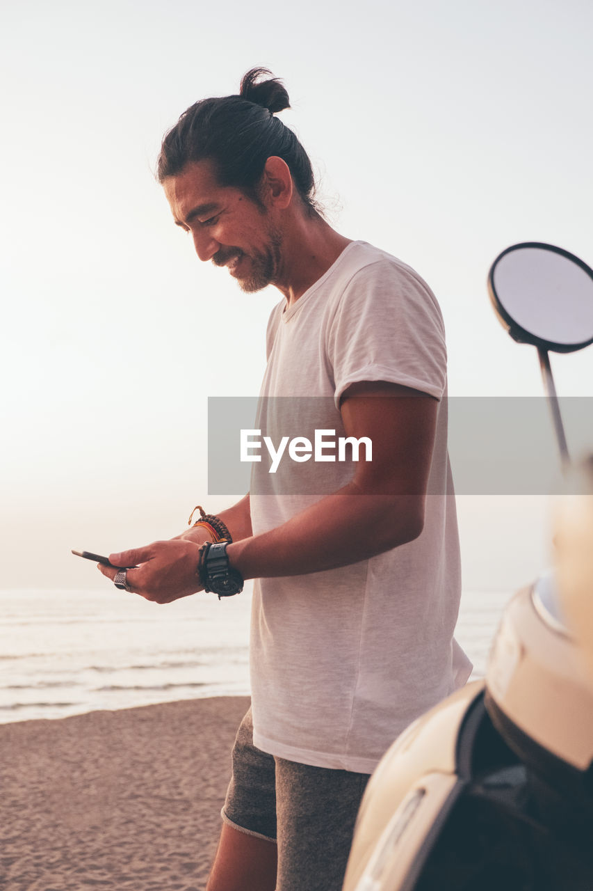 Side view of positive modern sporty male in t shirt and shorts standing next to bike and using mobile phone at seaside at sunset time