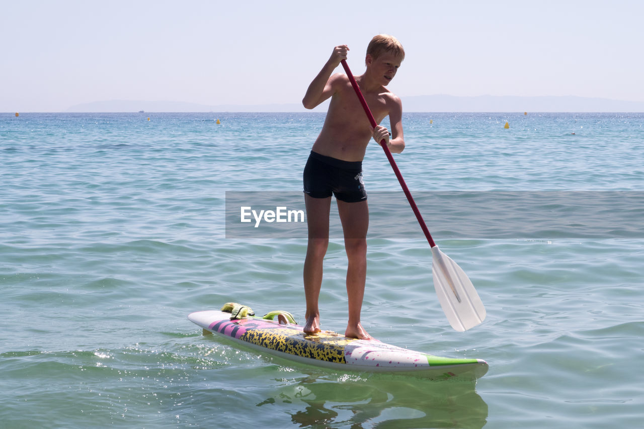 Shirtless teenage boy paddleboarding on sea