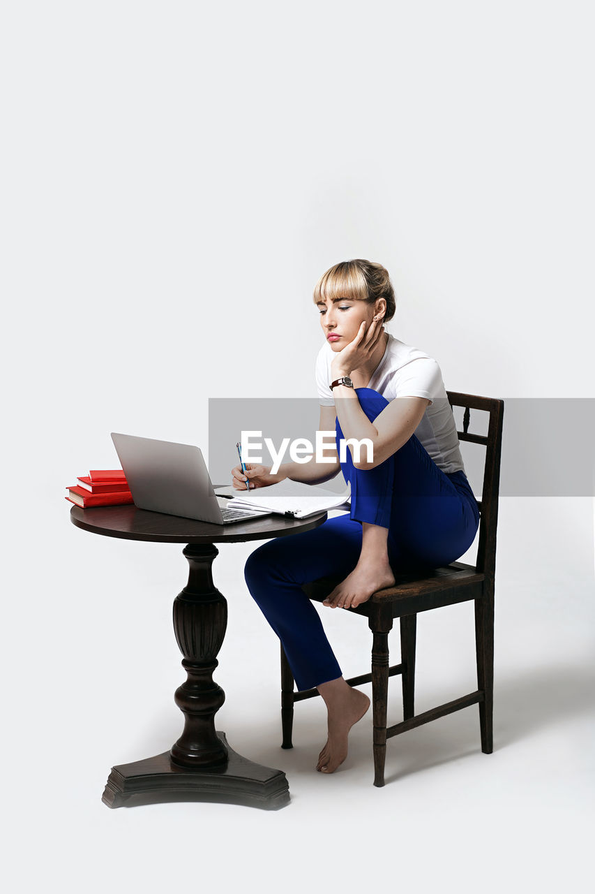 portrait of smiling young woman using laptop while sitting on chair against white background