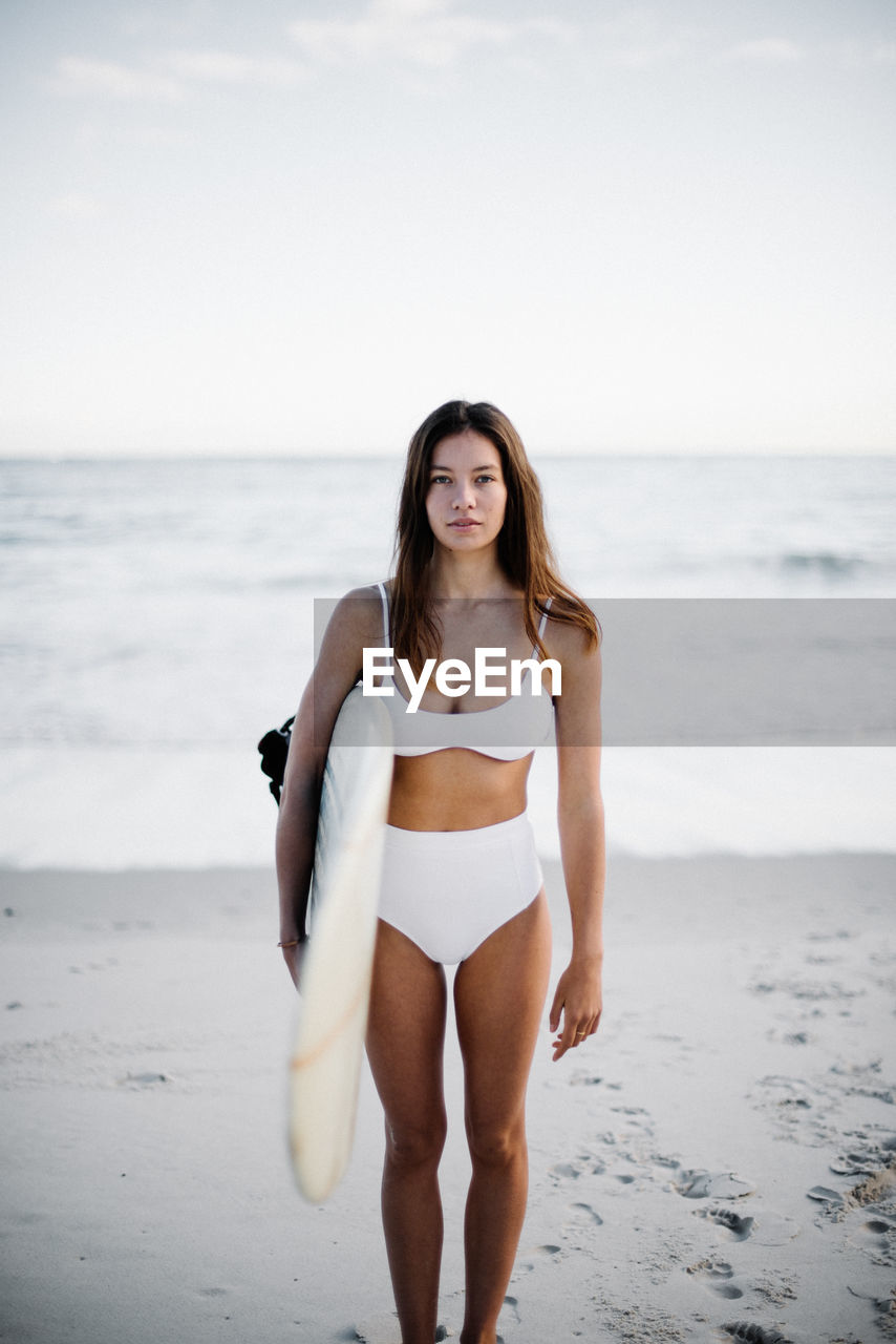 Portrait of young bikini woman with surfboard standing at beach