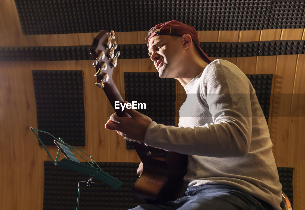 Caucasian man sings song, playing guitar, holding musical instrument in hands, sitting in chair in