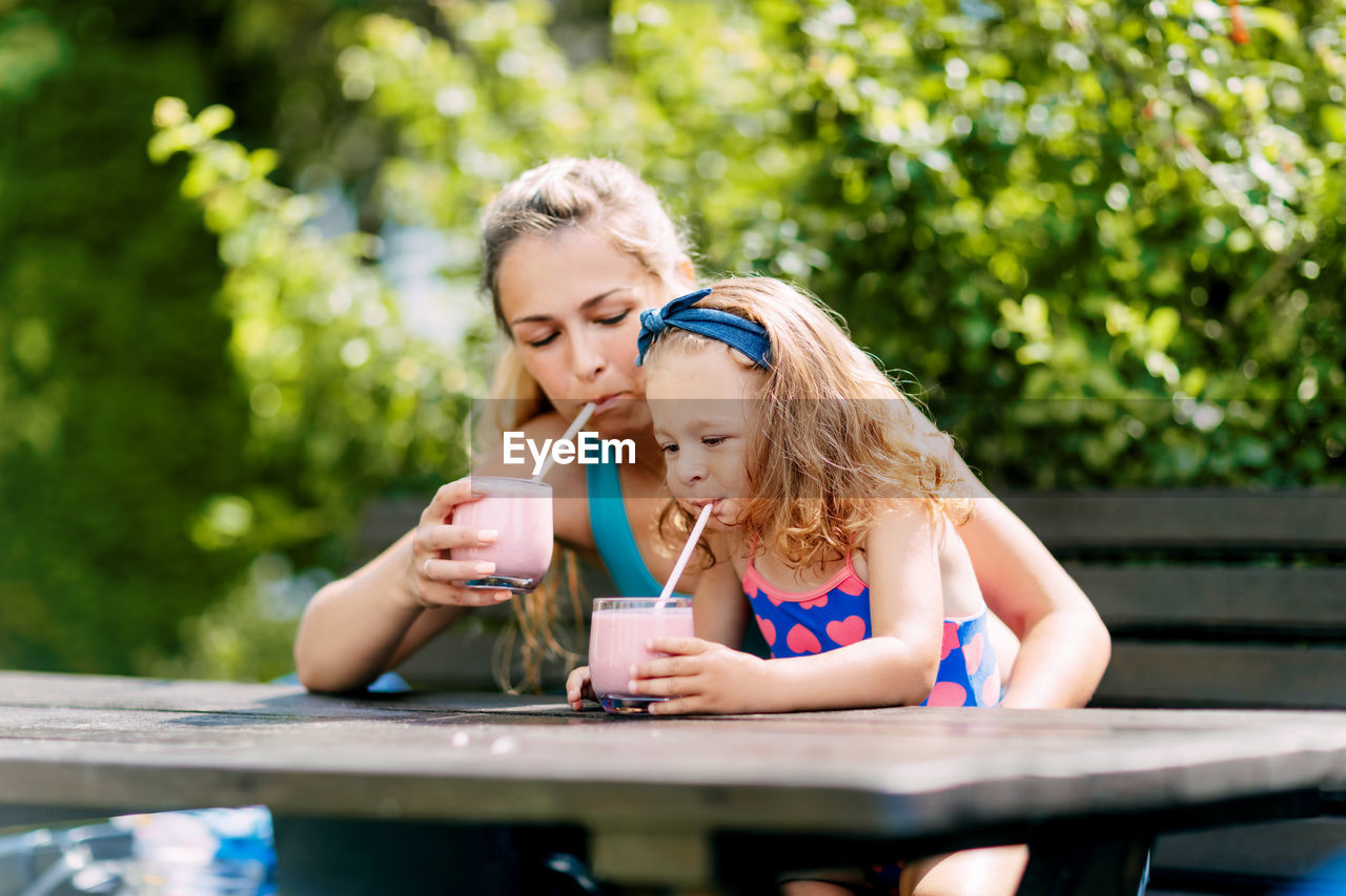 A beautiful girl and her little daughter drink a refreshing smoothie while sitting on a bench 