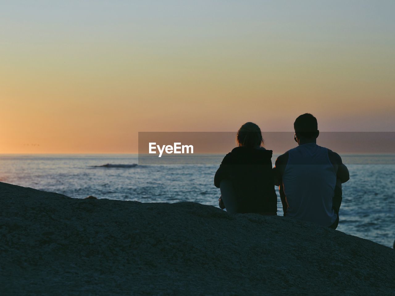 Couple sitting on cliff in front of sea during sunset