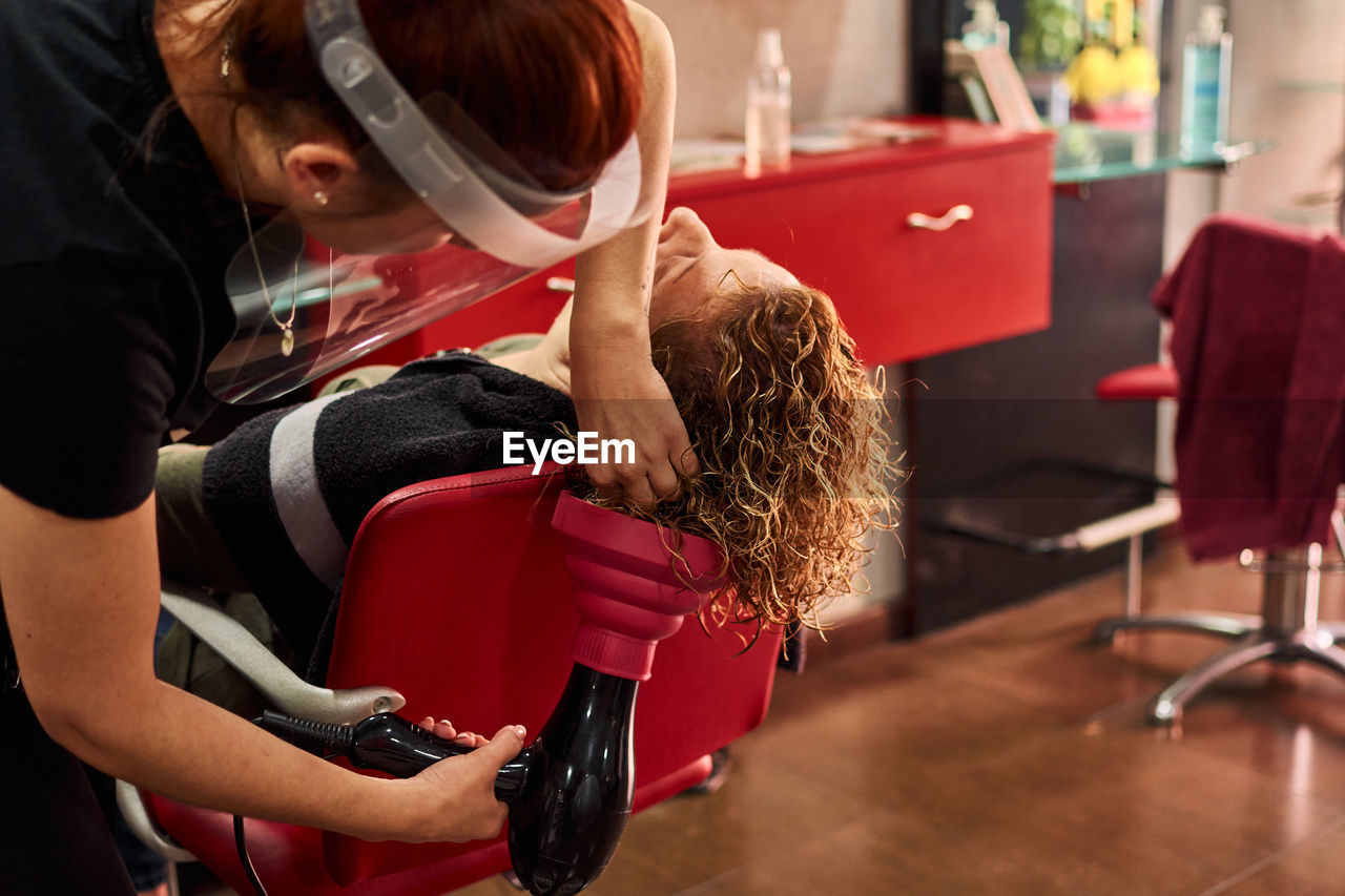 Hairdresser with a face shield dries her client's hair in her salon