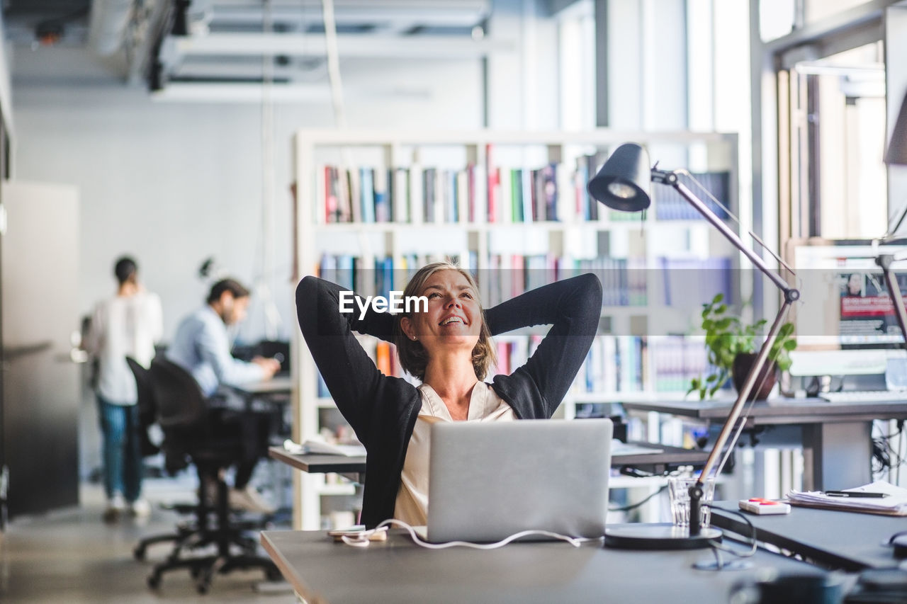 Mature businesswoman sitting with hands behind head at desk in office