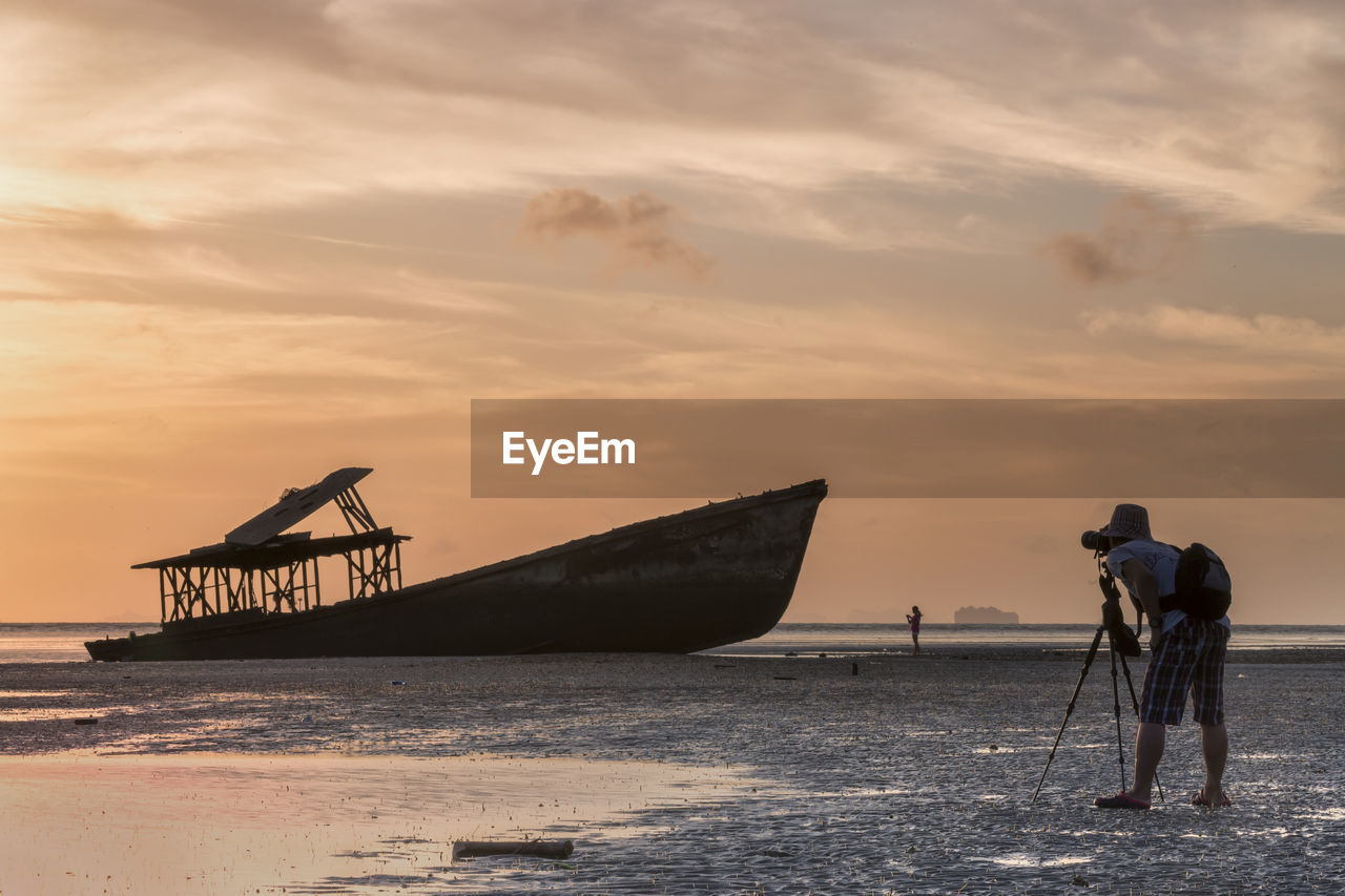 Side view of man photographing abandoned boat at beach against cloudy sky during sunset