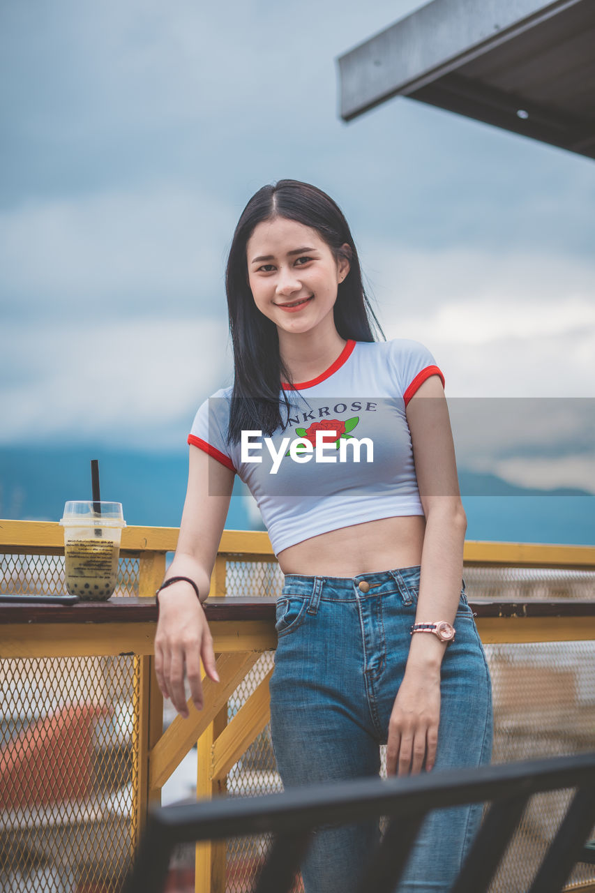 Portrait of smiling young woman standing against cloudy sky in city