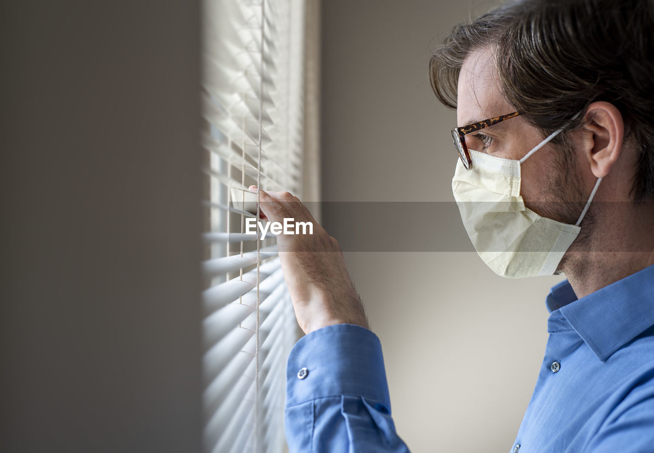 Side profile of man wearing respirator face mask looking out window, eye glasses.