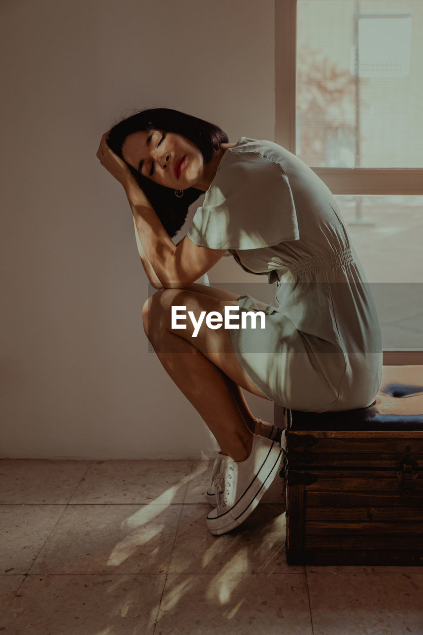 Side view of tender female leaning on hands while sitting with closed eyes on stool in room lit by sunlight