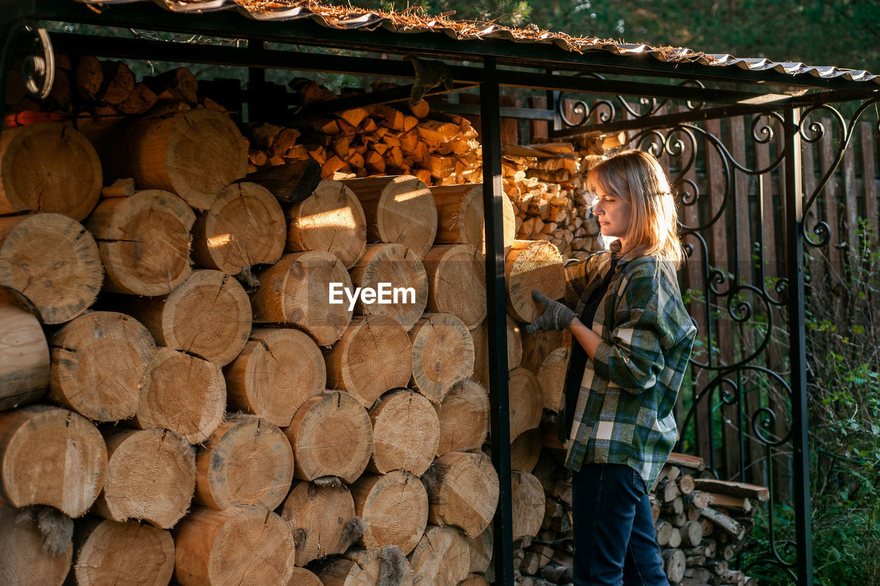 A beautiful blonde woman in a plaid shirt collects wood from a wood-burner. everyday rural life.