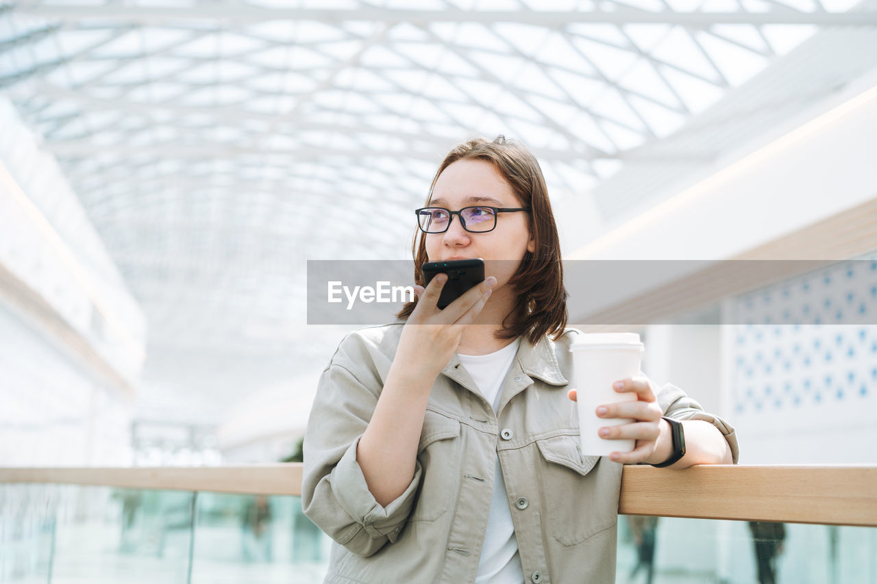 Young teenager girl in glasses using mobile phone sent voice message with coffee at shopping mall