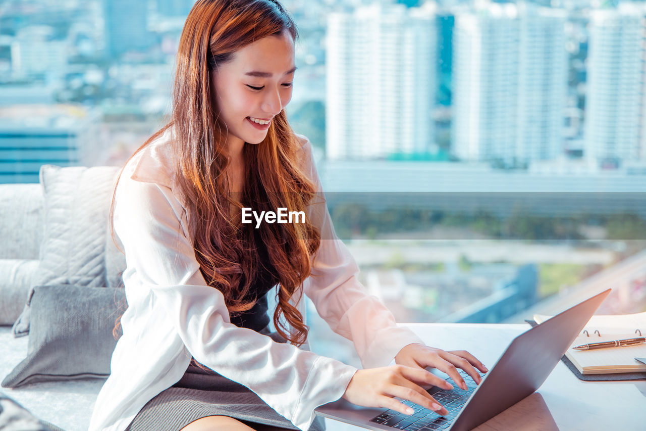 YOUNG WOMAN LOOKING AWAY WHILE SITTING ON MOBILE PHONE IN OFFICE