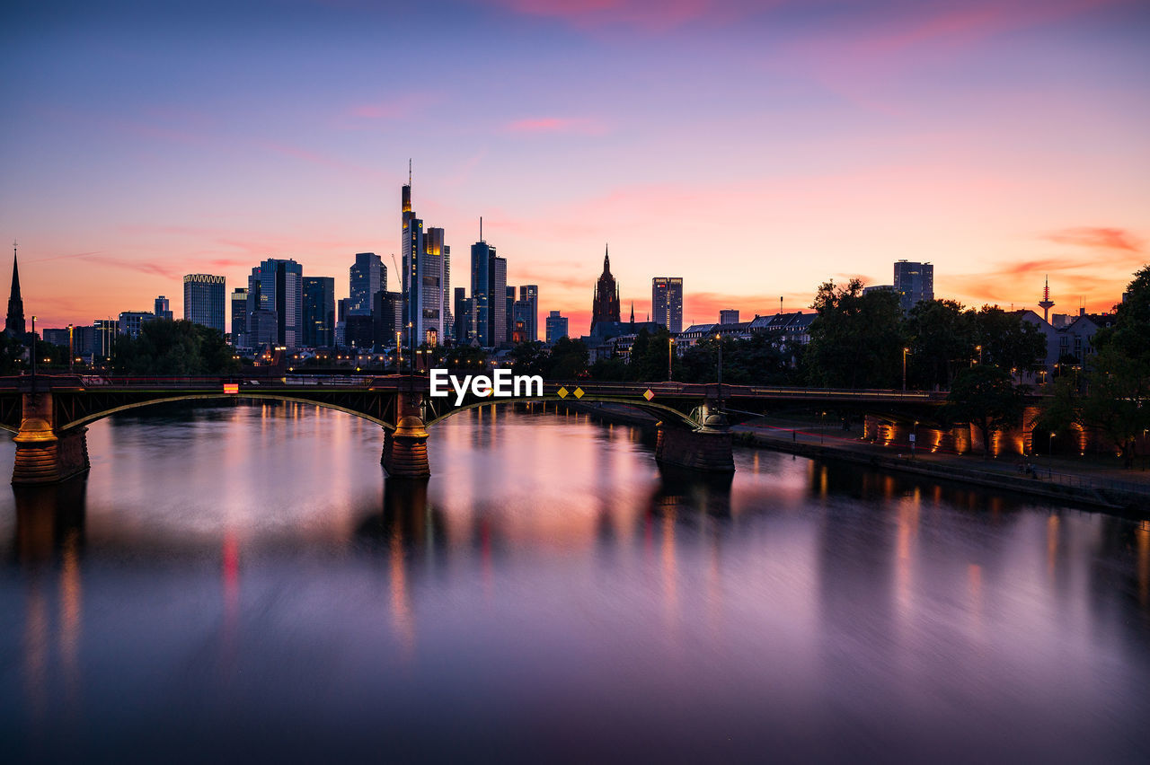 Scenic view of river by buildings against sky during sunset