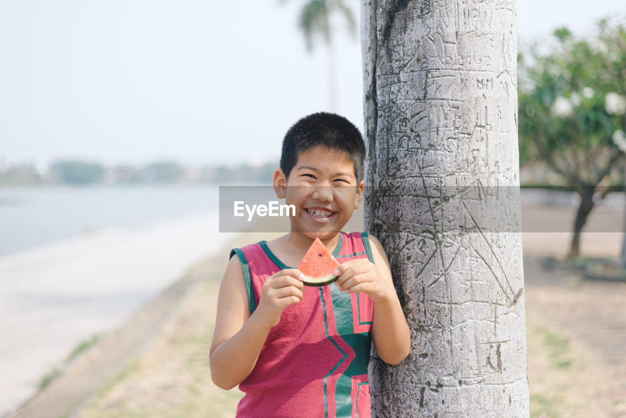 Portrait of smiling boy eating watermelon by tree