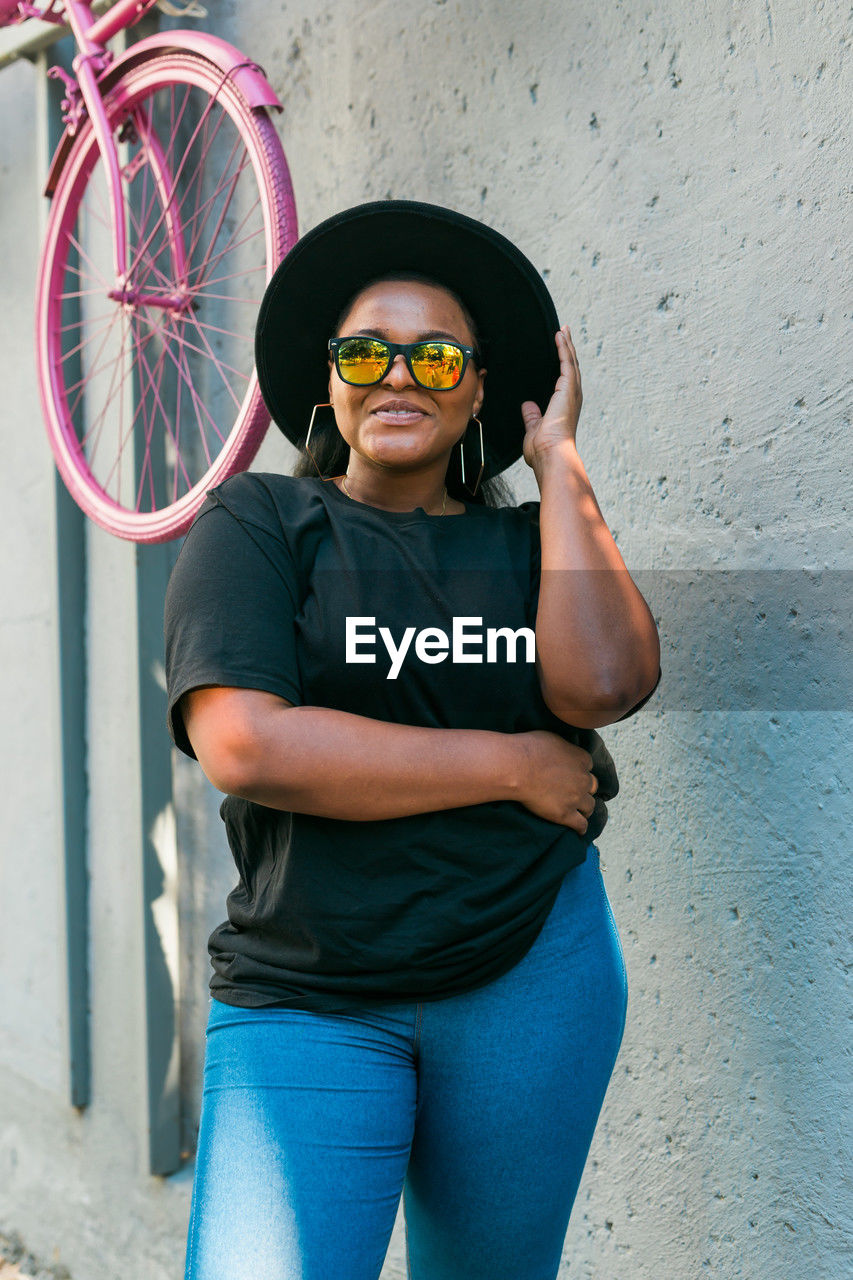 blue, bicycle, one person, glasses, adult, three quarter length, portrait, fashion, standing, sunglasses, looking at camera, wall - building feature, front view, casual clothing, young adult, architecture, clothing, lifestyles, day, eyeglasses, cool attitude, city, women, smiling, person, outdoors, red, leisure activity, men, black, vehicle, human face, fun