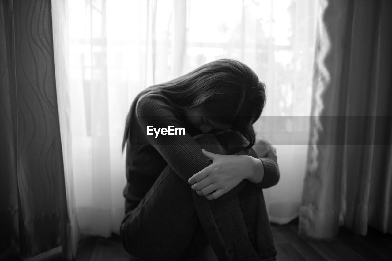 Depressed woman sitting against window at home